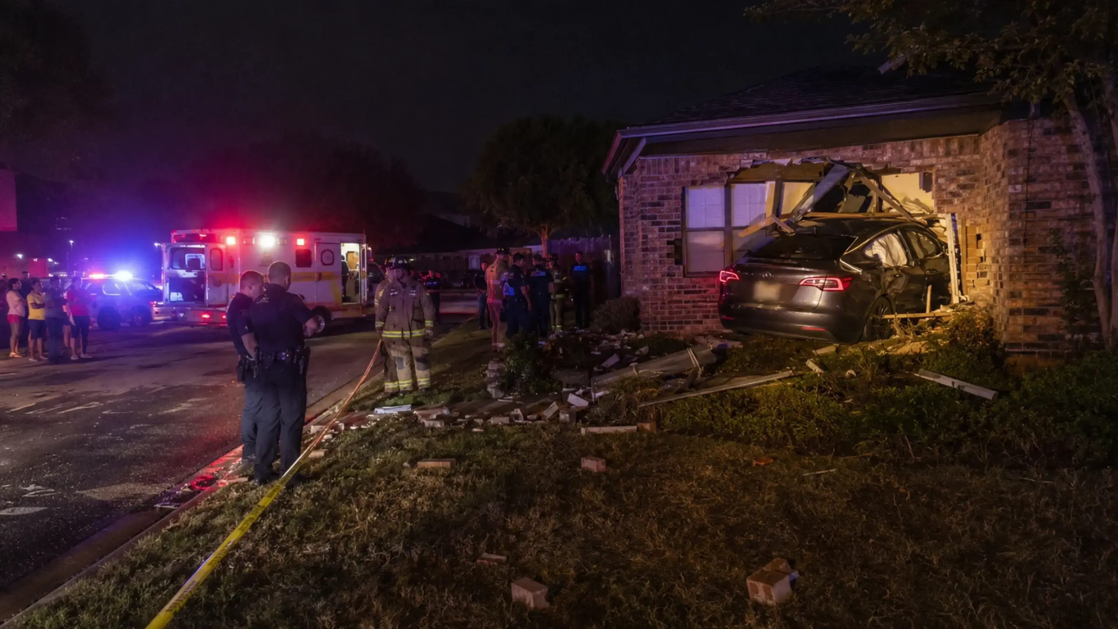Accidente nocturno en una zona residencial: un coche oscuro empotrado en la fachada de una casa, con gran destrozo en el muro, es atendido por policías, bomberos y una ambulancia bajo luces de emergencia.
